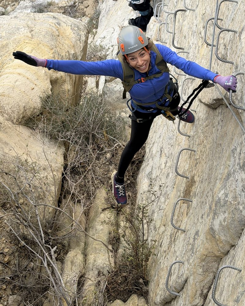 Elvia escalando una pared de roca con arnés y casco, sonriendo con los brazos abiertos